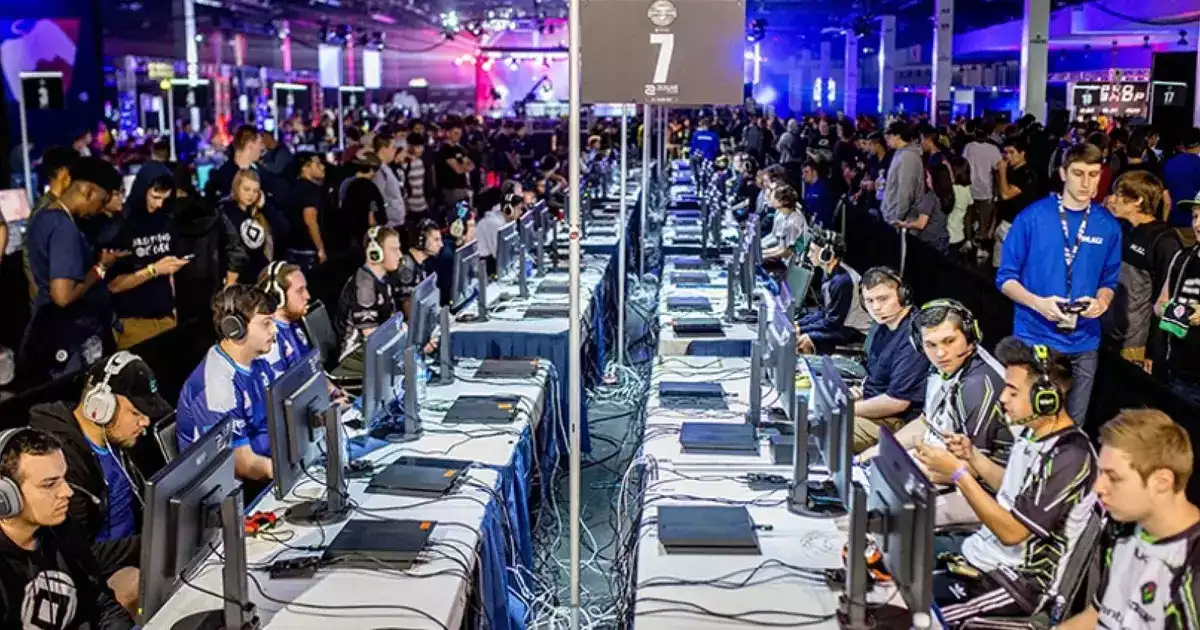 Image shows teams lined in up a row od desks set up and ready to compete. Crowns of fans and staff are stood behind the players